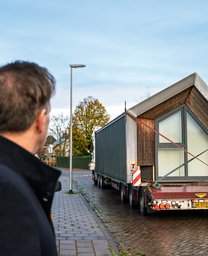 Rechts zie je een vrachtwagen met daarop het bovenste deel van de Heijmans ONE dat verplaatst wordt. Links zie je projectmanager Martijn Ras