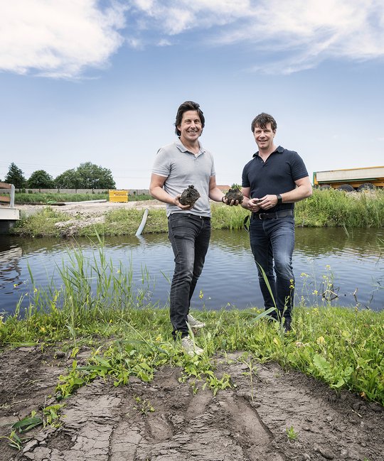 Lodewijk Hoekstra en een andere man staan bij een sloot en hebben modder in hun handen.